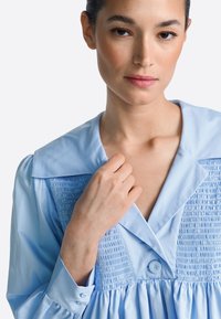 Vestido azul claro con cuello, cuerpo fruncido y detalle de botones. Mangas largas con botones en los puños, textura suave.