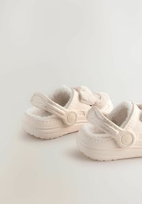 Chaussons beige clair avec doublure moelleuse, sangle ajustable avec accents métalliques, semelle texturée et forme arrondie. Matériaux doux partout.