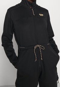 Černý teplákový komplet s vysokým límcem, předním zipem a dvěma kapsami na zip. Má stažení v pase pomocí šňůrky s texturovaným akcentem.