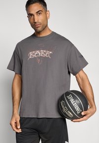 Szary bawełniany t-shirt z nadrukiem przedstawiającym słowo "Kobe" oraz skrzydła anioła, zestawiony z czarnymi spodenkami do koszykówki i trzymający piłkę do koszykówki.