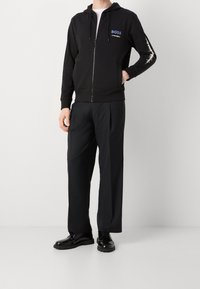 Felpa nera con zip e logo "BOSS" sul petto, abbinata a pantaloni neri a vestibilità ampia e scarpe nere lucide. Presenta cappuccio e polsini a costine.