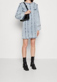 Džínové šaty v světle modré barvě s vertikálním štepováním, dlouhými rukávy, volnějším střihem a rozcupovaným lemem. Doplněné černými kotníkovými botami a malou kabelkou.