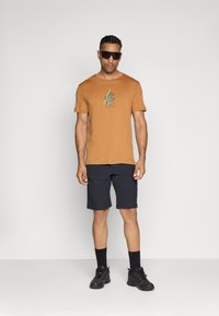 Brun t-skjorte med korte ermer og grafisk trykk, sammen med marineblå shorts. Sorte sokker og fottøy fullfører antrekket. Enkel, sporty design.
