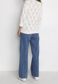 Valkoinen neulepaita, jossa on timanttimallinen tekstuuri, yhdistettynä löysiin sinisiin farkkuihin ja valkoisiin kenkiin.