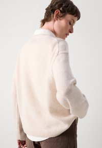 Beige strikkegenser med rund hals, ribbet søm og lange ermer, sett fra baksiden mot en nøytral bakgrunn.