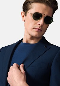 Granatowa marynarka o subtelnej teksturze i klapach w kształcie notcha, zestawiona z jednolitą granatową koszulą z okrągłym dekoltem. Okrągłe okulary przeciwsłoneczne z ciemnymi szkłami.