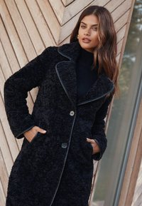 Palton exterior negru, pufos, cu guler tip șal, închidere frontală cu nasturi și buzunare laterale, detaliat cu margini pipate negre.
