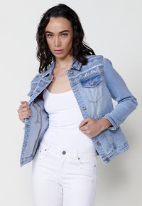 Világoskék farmerdzseki díszvarrással és mintás vállrésszel. Két gombos zsebe van elöl, és egy fehér trikót visel alatta.