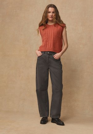 Jeune femme aux longs cheveux ondulés portant un haut tricoté orange sans manches, un jean ample gris foncé et des mocassins noirs, mains dans les poches.