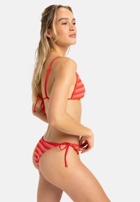 Bikini de rayas rojas y naranjas con lazos ajustables; parte superior triangular y parte inferior de corte bajo que destacan una textura suave y un diseño minimalista.