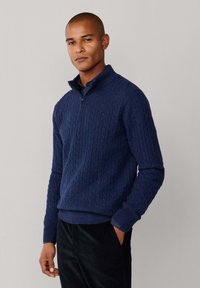 Granatowy sweter z wełny z wysokim kołnierzem i ćwierć zamkiem. Posiada teksturowany wzór w warkocze oraz ściągacze na rękawach, noszony na ciemnej koszuli.