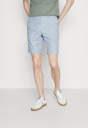 Man in lichtblauwe shorts, groene shirt ingestopt, witte sneakers met gumzolen, staand met één hand in de zak tegen een effen achtergrond.