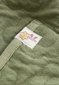 Grön quiltad tyg med ett texturerat mönster; en sydd etikett med en design av en gul blomma med rosa detaljer.