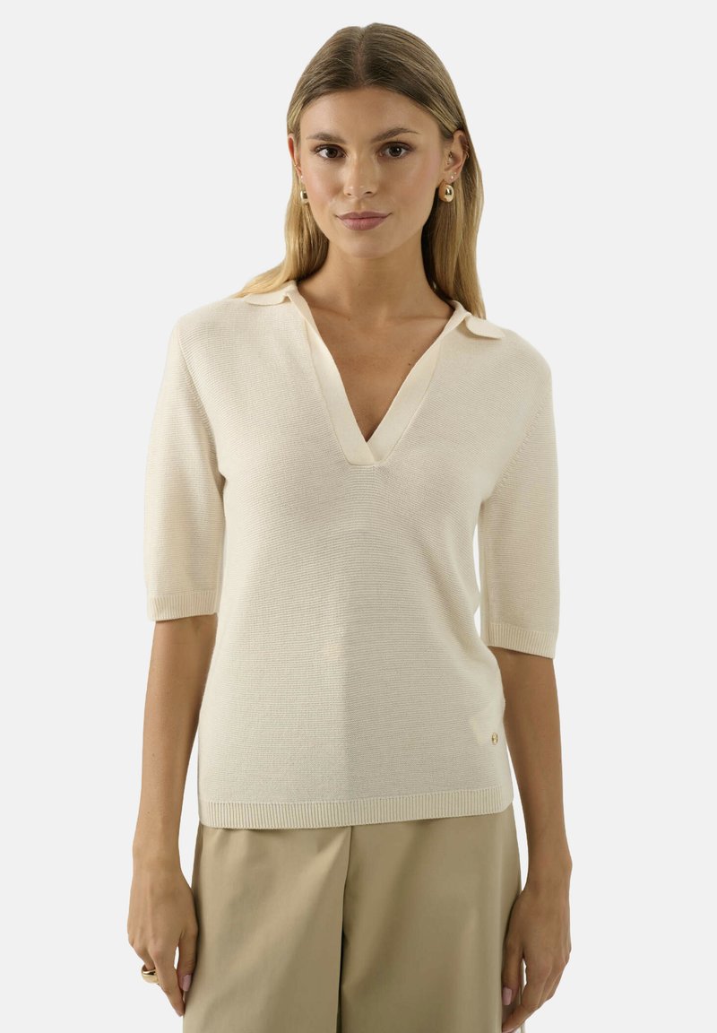 Donna che indossa un top a maniche corte di maglia color crema con scollatura a V profonda e pantaloni beige, in piedi contro uno sfondo chiaro e semplice.