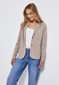 Ljusbeige blazern med fickklapp, två framfickor och enknappsstängning, som bärs över en vit topp och blåjeans.