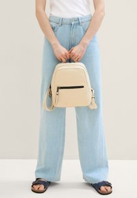 Zaino in finta pelle beige con una tasca frontale con zip, forma arrotondata e spallacci regolabili, abbinato a jeans a gamba larga di un blu chiaro.