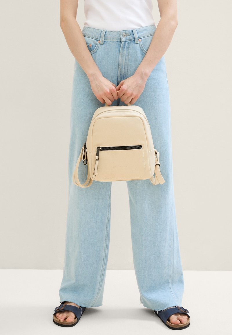 Zaino in finta pelle beige con una tasca frontale con zip, forma arrotondata e spallacci regolabili, abbinato a jeans a gamba larga di un blu chiaro.