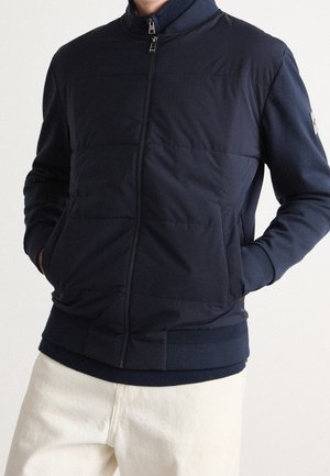 Mężczyzna w granatowej pikowanej kurtce zapinanej na zamek z prążkowanym kołnierzem, mankietami i dołem, w połączeniu z jasnobeżowymi spodniami, z rękami w kieszeniach.