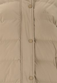 Beige puffertakki, jossa on vaakasuorat ompeleet, pyöreät napit ja pieni logo painettuna yläpuolelle. Sileä, hieman kiiltävä kangas.