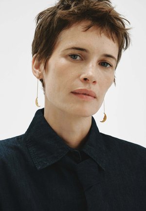 Mujer con cabello corto y castaño, llevando pendientes dorados con forma de luna creciente y una camisa oscura con cuello, mirando ligeramente fuera de cámara.