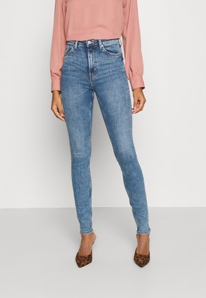 Kvinde iført højttaljede lyseblå skinny jeans, lyserød bluse med lange ærmer og leopardmønstrede spidse hæle, stående mod en ensfarvet baggrund.