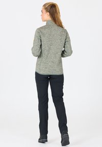 Fleecejacka i grön melerad tyg, hög krage och åtsittande design, matchad med svarta byxor. Slät textur med minimala detaljer.