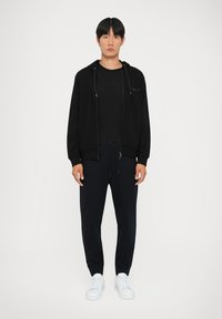 Svart zip-up hettegenser over en svart t-skjorte, kombinert med mørke joggebukser og hvite sneakers. Har en lomme foran og snor i livet.