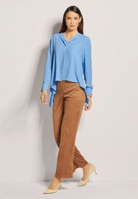 Blaues Langarmbluse mit V-Ausschnitt und asymmetrischem Saum, kombiniert mit braunen weit geschnittenen Hosen und beigen High-Heels.