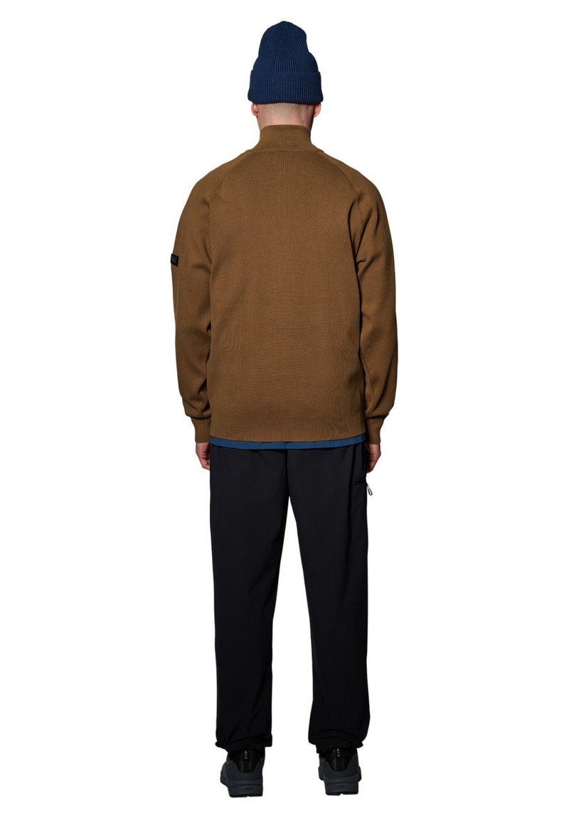 Brauner, strukturierter Fleece-Pullover mit hohem Kragen. Kombiniert mit dunklen Hosen und schwarzen Schuhen. Getragen mit einer blauen Strickmütze.