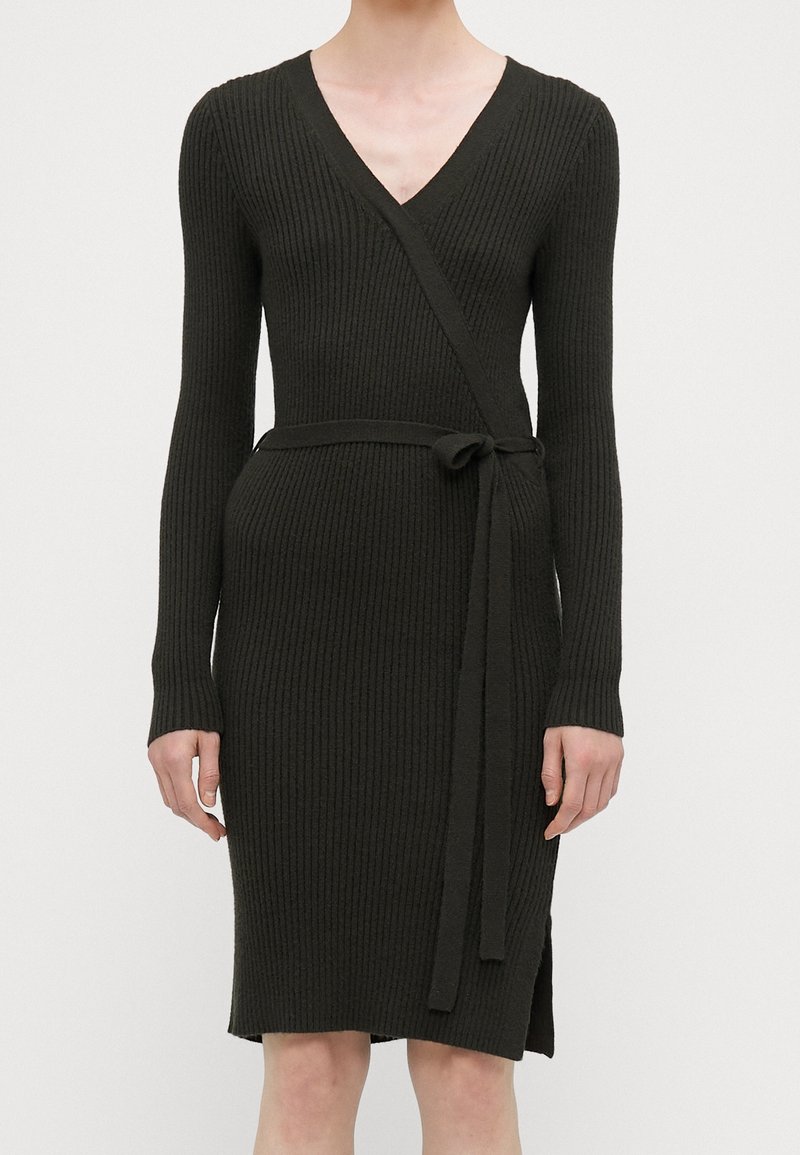 Robe portefeuille en maille côtelée noire avec manches longues et ceinture nouée à la taille, portée par une personne debout devant un fond clair uni.