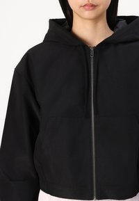Black zip-up jacket with a hood, featuring a front pocket and smooth texture. Simple design, highlighting a modern silhouette.