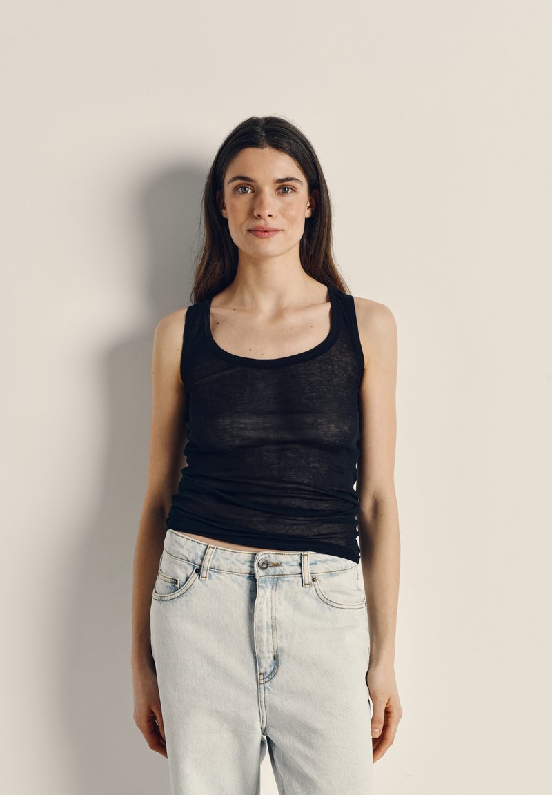 Young woman with long dark hair wearing a black sleeveless top and light blue jeans, standing against a plain beige wall.
