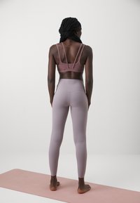 Vista posteriore di una persona che indossa un reggiseno sportivo aderente di colore malva con spalline regolabili e leggings a vita alta di un malva chiaro, su un tappetino da yoga rosa.