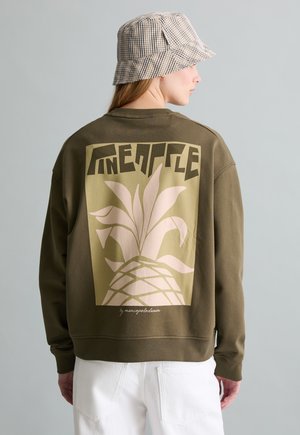 Persona che indossa una felpa verde oliva con una grande grafica di ananas beige chiaro e testo sul retro, abbinata a pantaloni bianchi e un cappello a secchiello a quadri.
