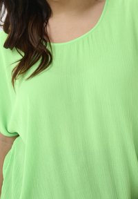 Femme portant un haut vert clair ample et texturé avec un col rond, cheveux ondulés foncés partiellement visibles sur l'épaule.