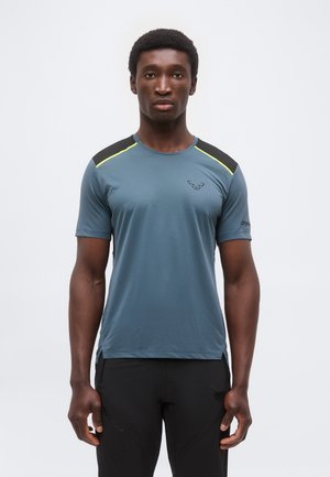 Egy férfi áll, előre néz, kék sportos pólót visel fekete és sárga válldíszítéssel, valamint fekete nadrágot, egyszerű fehér háttér előtt.