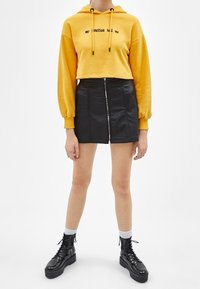 Persona que lleva un cropped amarillo con texto, falda mini negra con cremallera frontal, calcetines blancos y botas de plataforma negras con cordones.
