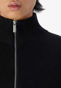 Svart ribbad stickad zip-up tröja med hög krage och silverfärgad dragkedja, som visar en texturerad tyg.