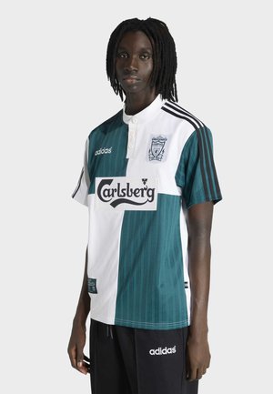 Young person with dark skin and black dreadlocks wearing a half white, half green Liverpool football jersey with Carlsberg logo.