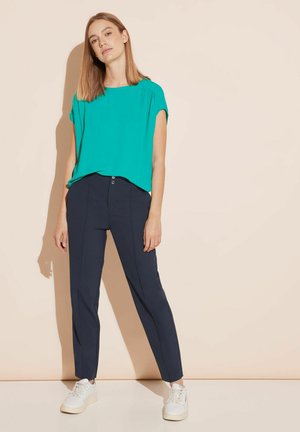 Haut turquoise à manches courtes assorti à un pantalon noir marine ajusté. Le mannequin porte des baskets blanches. Tissu lisse et coupe décontractée.