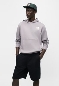 Νεαρός άνδρας με ανοιχτό γκρι φούτερ North Face, μαύρο σορτς και πράσινο καπέλο μπέιζμπολ, στέκεται με τα χέρια στις τσέπες κοιτάζοντας αριστερά.