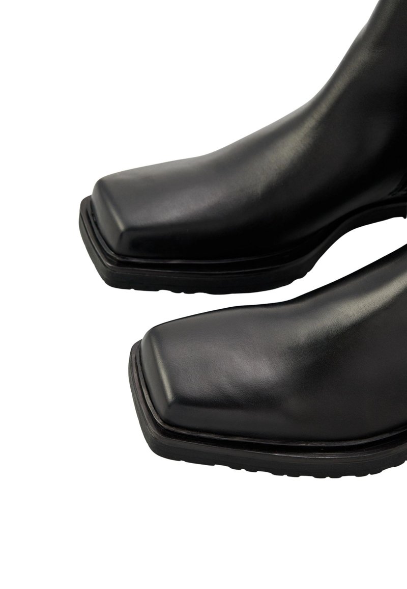 Stivali neri in pelle alla caviglia con punta quadrata, texture liscia, suola piatta in gomma e dettagli di cucitura minimi.