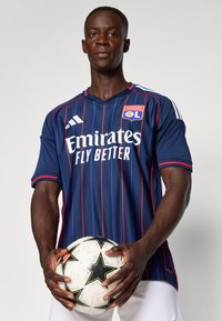 Tamsiai mėlyna futbolo marškinėliai su raudonomis juostomis, su Adidas logotipu, "Emirates" tekstu ir klubo simboliu; žaidėjas laiko baltą futbolo kamuolį su juodu staru.
