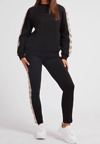 Conjunto de sudadera y pantalones negros. La sudadera tiene cuello redondo y detalle de logo; los pantalones cuentan con franjas laterales y un corte ajustado, combinados con zapatillas blancas.
