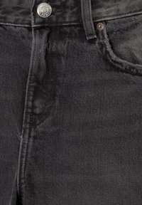 Gros plan sur un jean en denim noir montrant le bouton supérieur, la boucle de ceinture, la poche avant et le détail du rivet en métal.