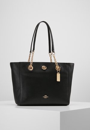 Zwarte leren totebag met gouden kettinghandvatten, draaivergrendeling en een klein hangend gouden label op een wit voetstuk.