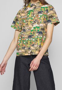 Košile s krátkým rukávem na knoflíky v vzorované tkanině s různými barevnými zvířaty a rostlinami na khaki pozadí.