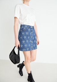 Person wearing a white t-shirt, blue patterned button-front denim skirt, black ankle boots, and holding a black handbag while walking.