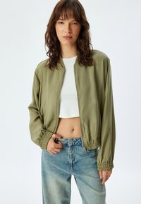 Veste bomber verte olive avec fermeture éclair à l'avant, poignets élastiqués et coupe décontractée, associée à un jean taille haute bleu clair.