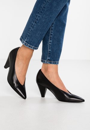 Stopy w czarnych skórzanych butach na średnim obcasie z ostrykiem, w zestawieniu z niebieskimi, podwiniętymi dżinsami na białym tle.